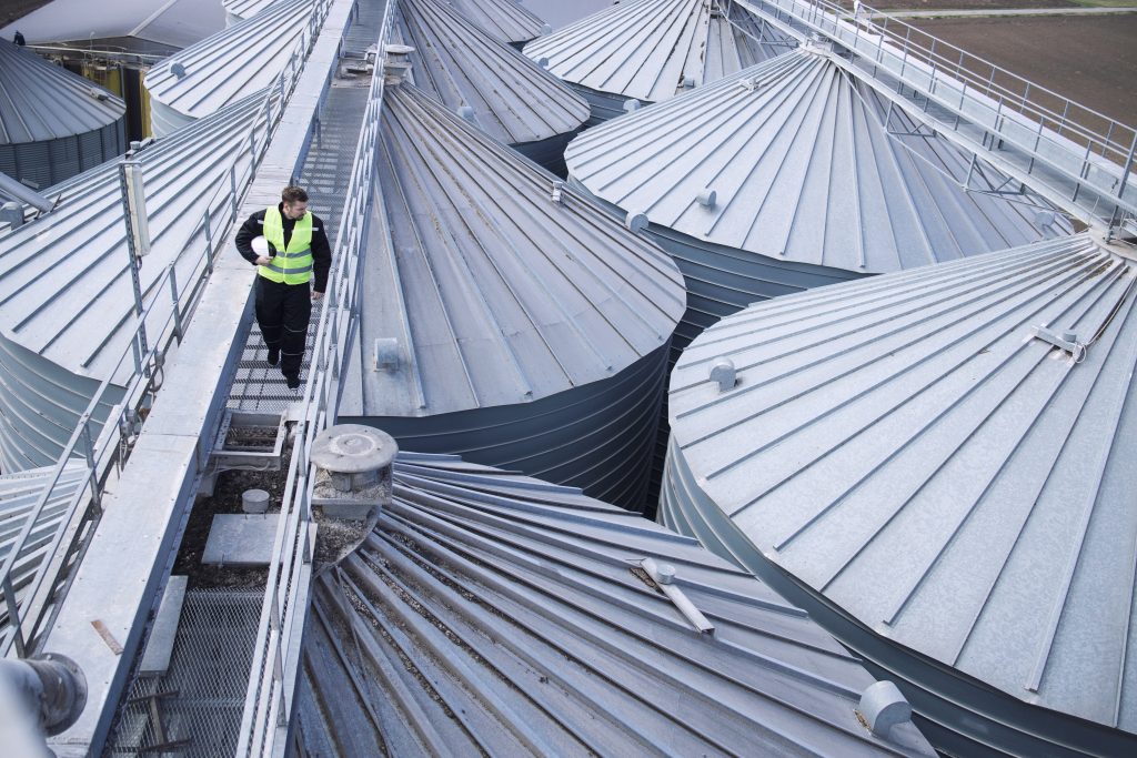 Factory worker walking on metal platform and doing visual inspection on industrial food storage tanks or silos.